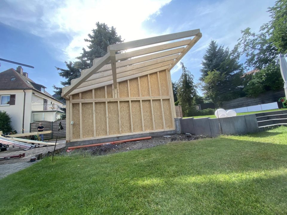 Der beplankte Giebel vom Garten aus gesehen, darüber das flach geneigte Pultdach mit seinen auskragenden Sparren vor hellem Himmel.