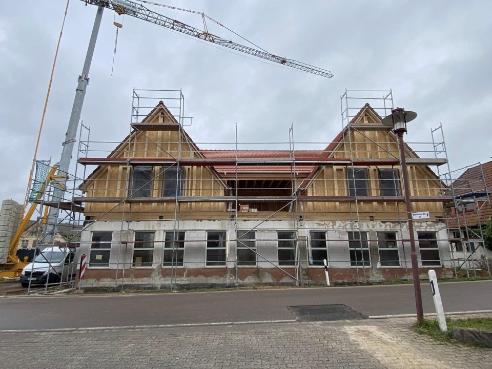 Straßenansicht: Das verputzte Erdgeschoss mit seiner langen Fensterreihe trägt das neue Obergeschoss in Holzrahmenbauweise mit zwei Zwerchgiebeln, dahinter ragt ein Kran auf.